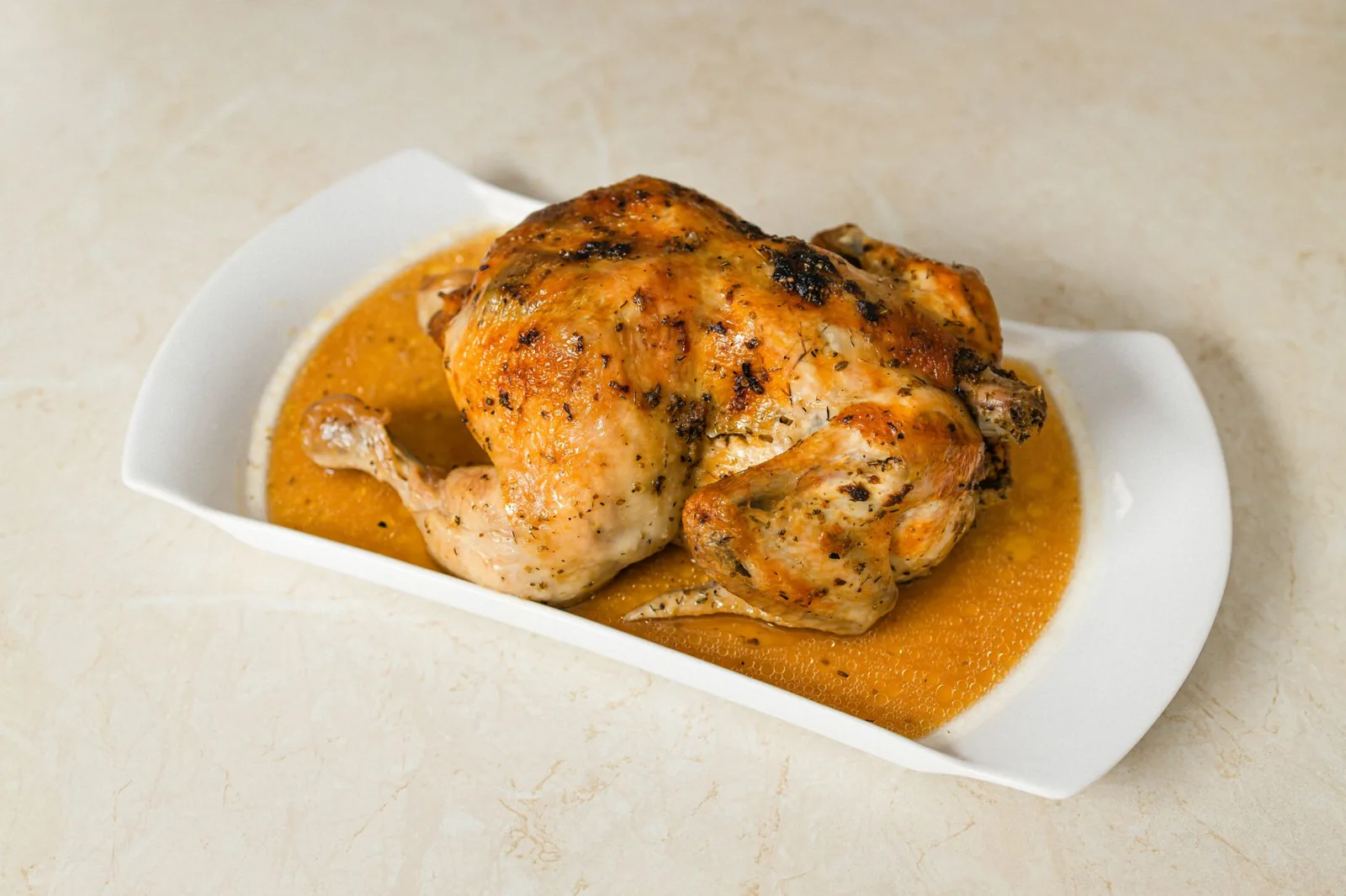 A whole roasted chicken sitting intact in its juices on a white serving dish, uncarved
