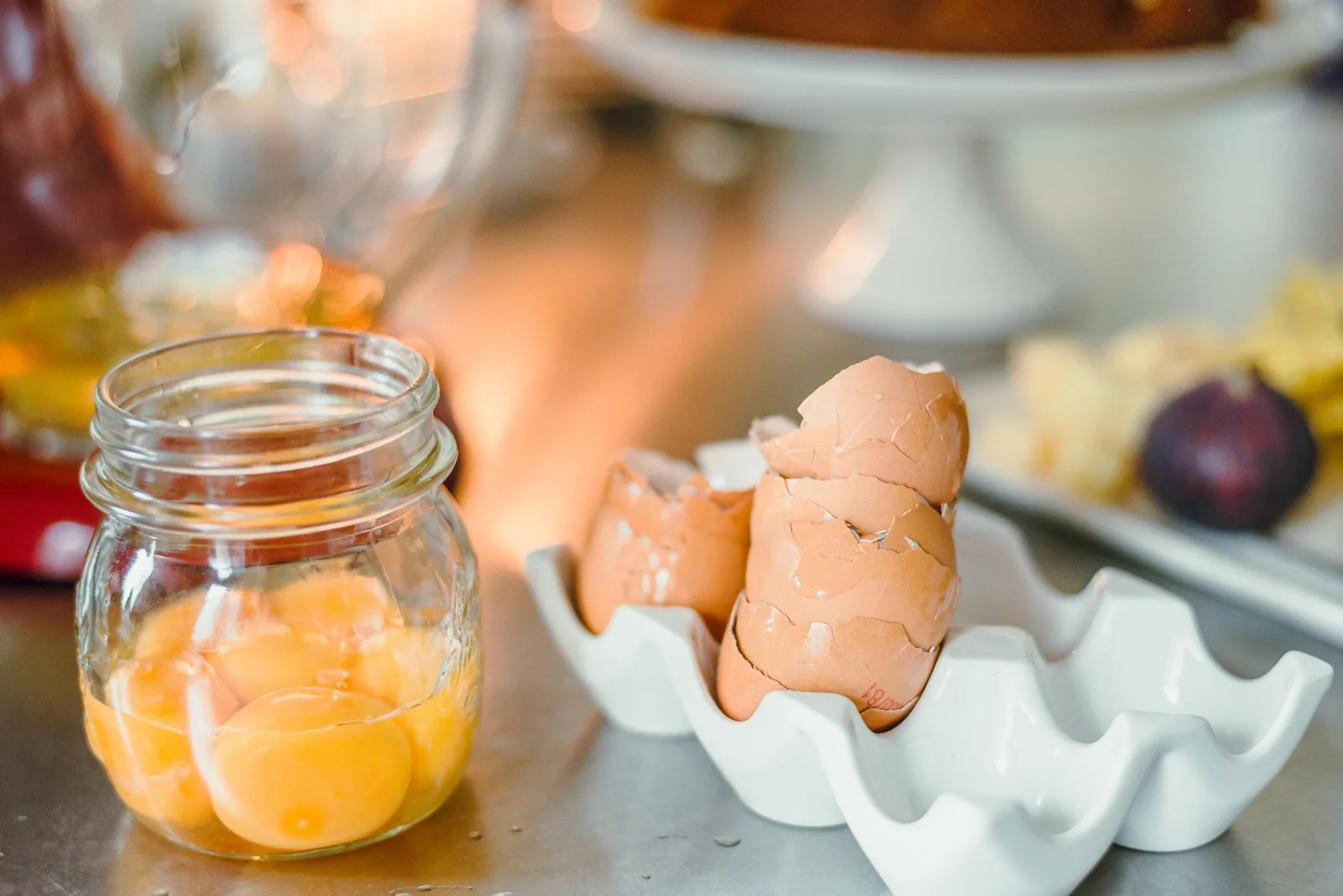 Raw egg yolks in a glass jar beside broken brown eggshells stacked in a white egg holder on a kitchen counter.