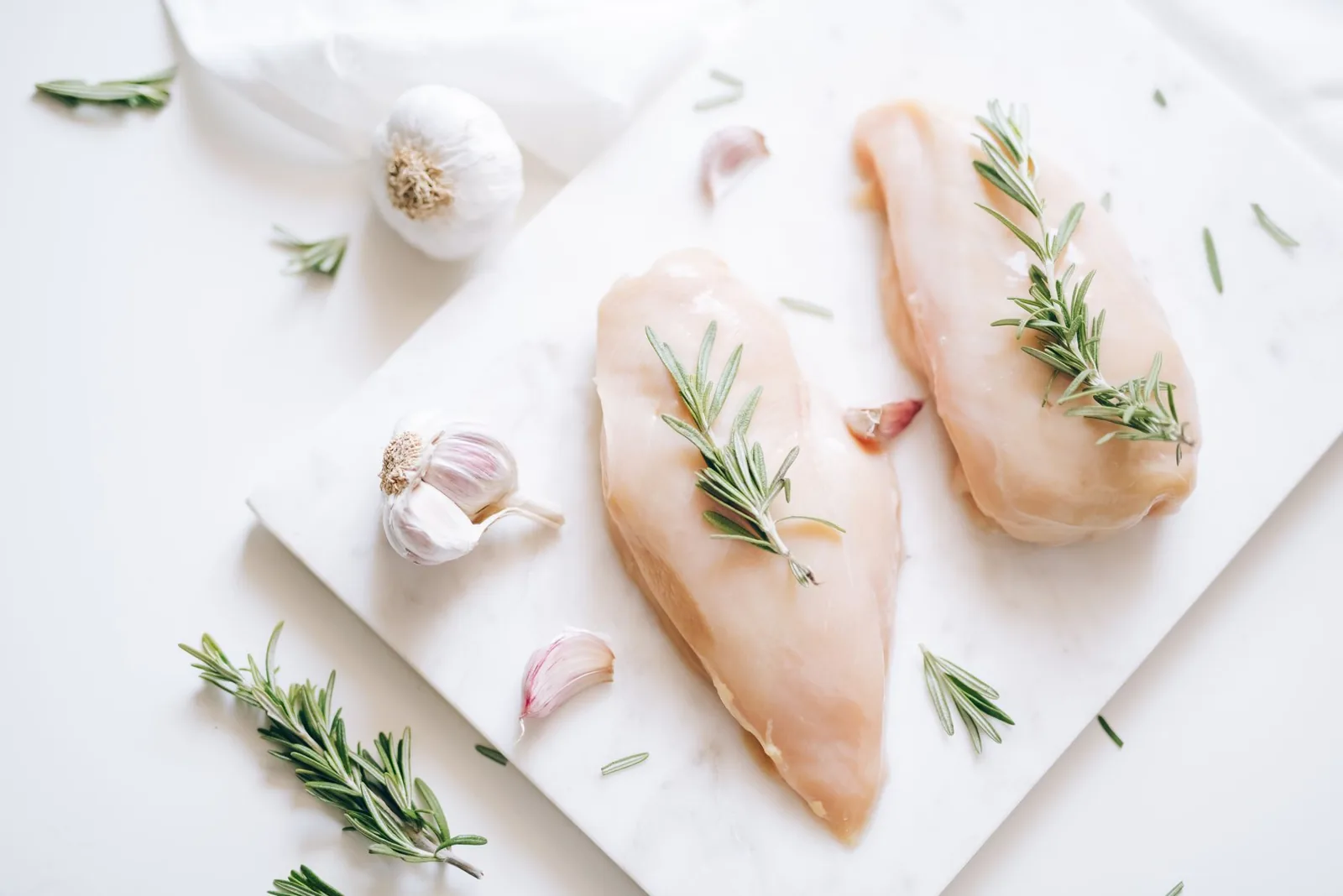Two raw chicken breasts on a marble board, their surface smooth and evenly pale pink with no visible white stripes, with rosemary sprigs and garlic cloves around them