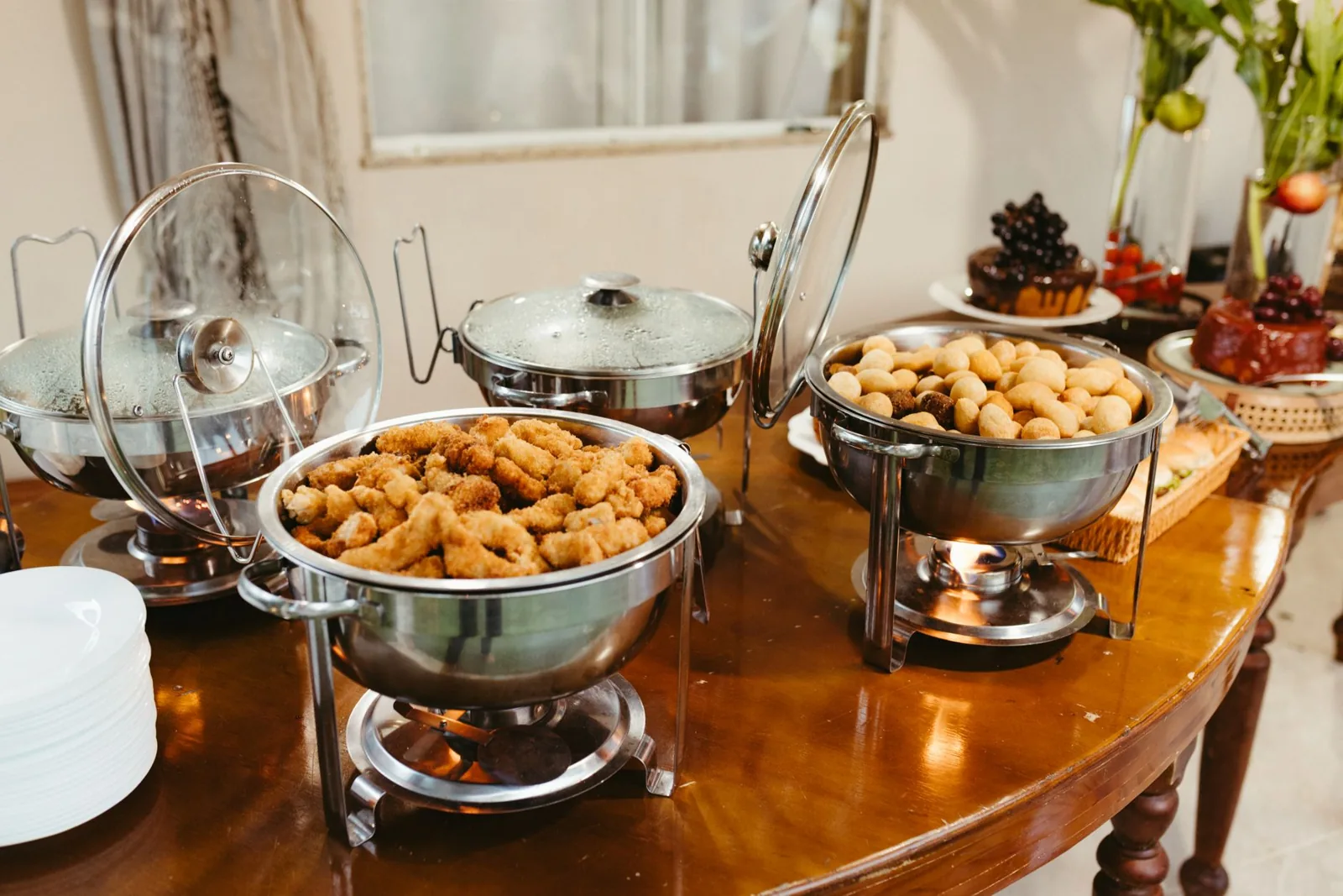 A wooden buffet table with three open chafing dishes, one of them filled with breaded fried chicken pieces, their glass lids propped up to one side.