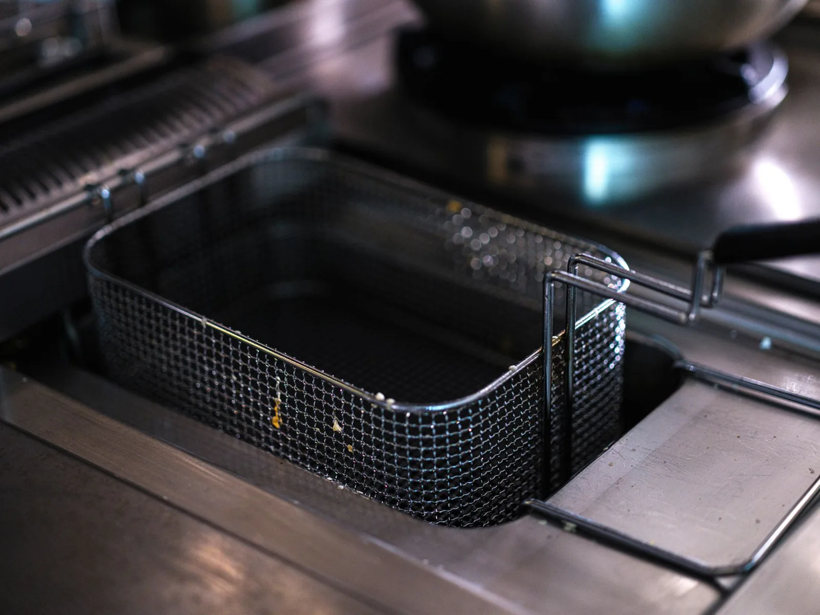 An empty metal frying basket sitting in a switched-off fryer in a commercial kitchen, a burner and a steel pan behind it, with nothing being cooked.